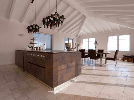 Kuchyňský ostrůvek s držákem na láhve vína na stropě, jídelní stůl a židle v pozadí na položeném travertinu Light s tromlovaným, nesrovnaným povrchem