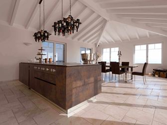 Kuchyňský ostrůvek s držákem na láhve vína na stropě, jídelní stůl a židle v pozadí na položeném travertinu Light s tromlovaným, nesrovnaným povrchem