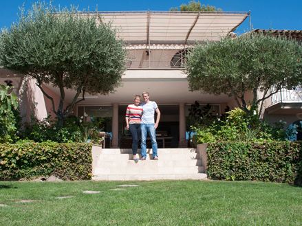 Pár stojící před hotelem s travertinovou podlahou, trávníkem v popředí a stromy vlevo a vpravo.