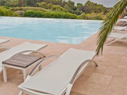 Bílé lehátko na travertinové podlaze u bazénu, zprava vyčnívá do obrazu palmová větev.