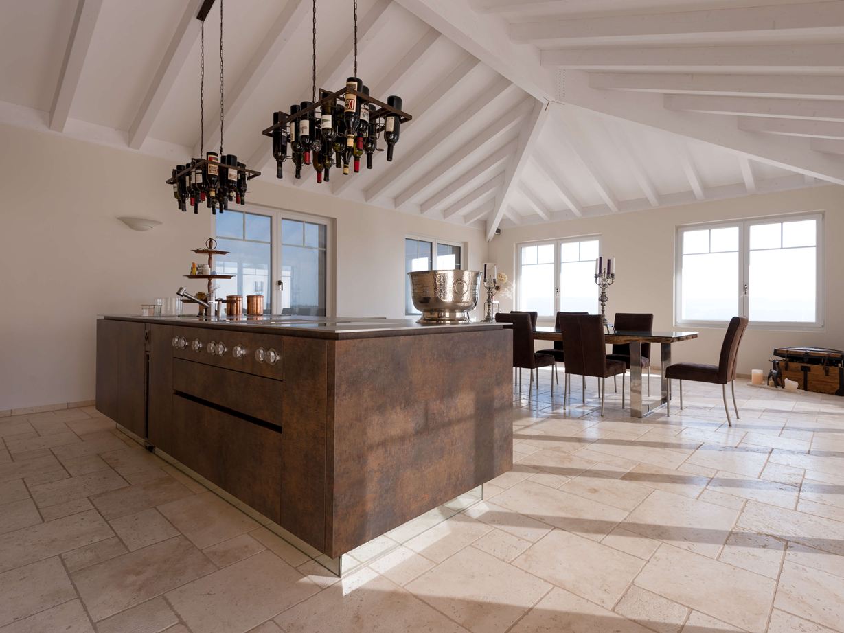 Kuchyňský ostrůvek s držákem na láhve vína na stropě, v pozadí jídelní stůl a židle na položeném travertinu Light s tromlovaným, nesrovnaným povrchem