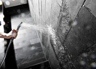 Ruka drží sprchovou hlavici u stěny s tmavými kamennými dlaždicemi.