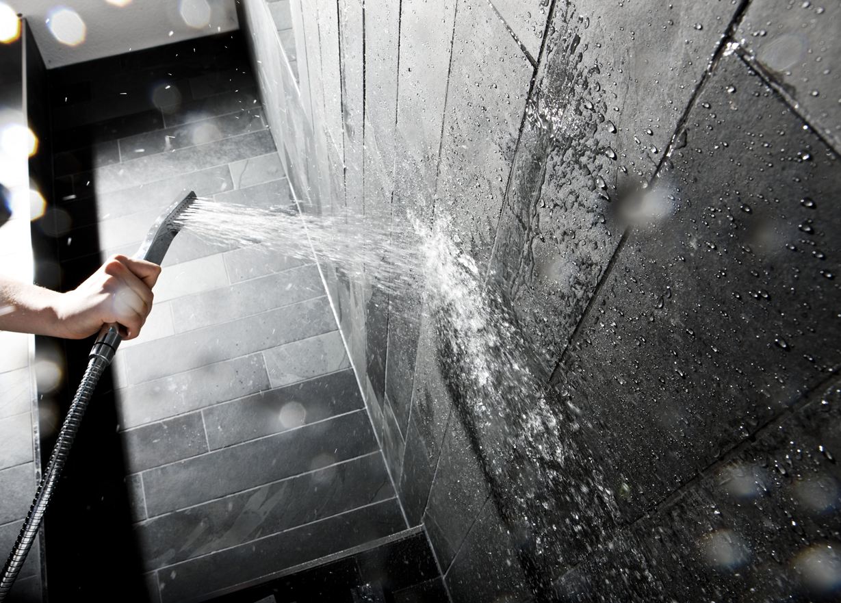 Ruka drží sprchovou hlavici u stěny s tmavými kamennými dlaždicemi.