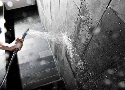 Ruka drží sprchovou hlavici u stěny s tmavými kamennými dlaždicemi.