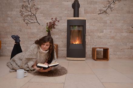 Žena leží na vápencových dlaždicích před stěnou s obložením z přírodního kamene a krbem a čte si knihu.